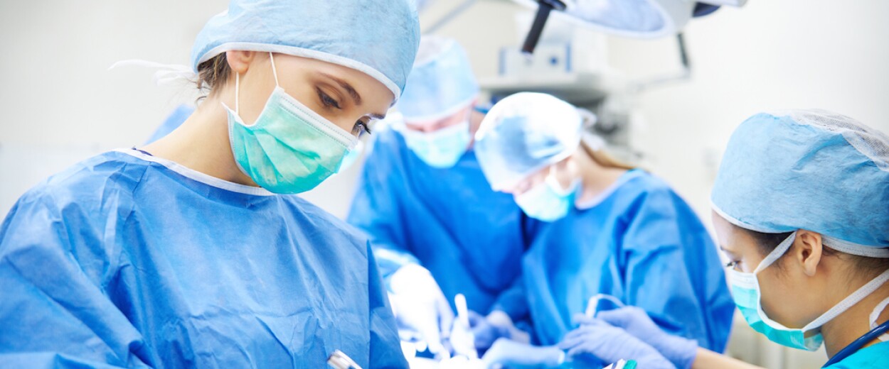 Female nurse in full scrubs and face mask marking notes on a clipboard in front of other medical personnel performing surgery