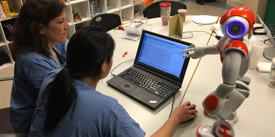 Two female nurses in scrubs looking at a laptop with a robot next to them