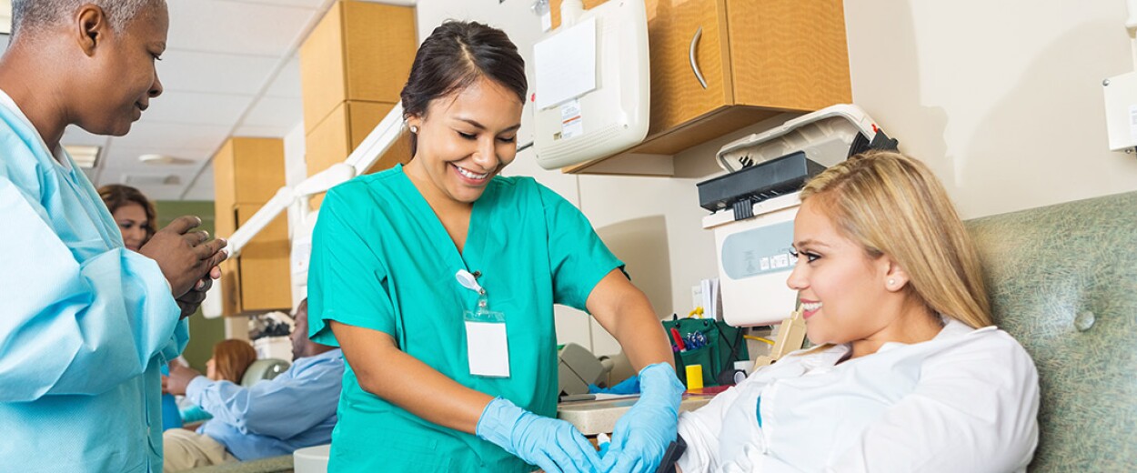 Female nurse in scrubs being observed taking blood from a female patient by another female nurse in scrubs