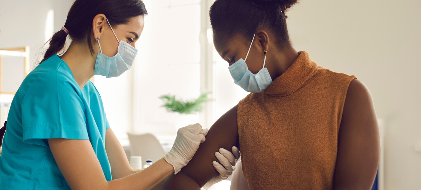 Female nurse in scrubs and face mask administering shot to female patient in face mask