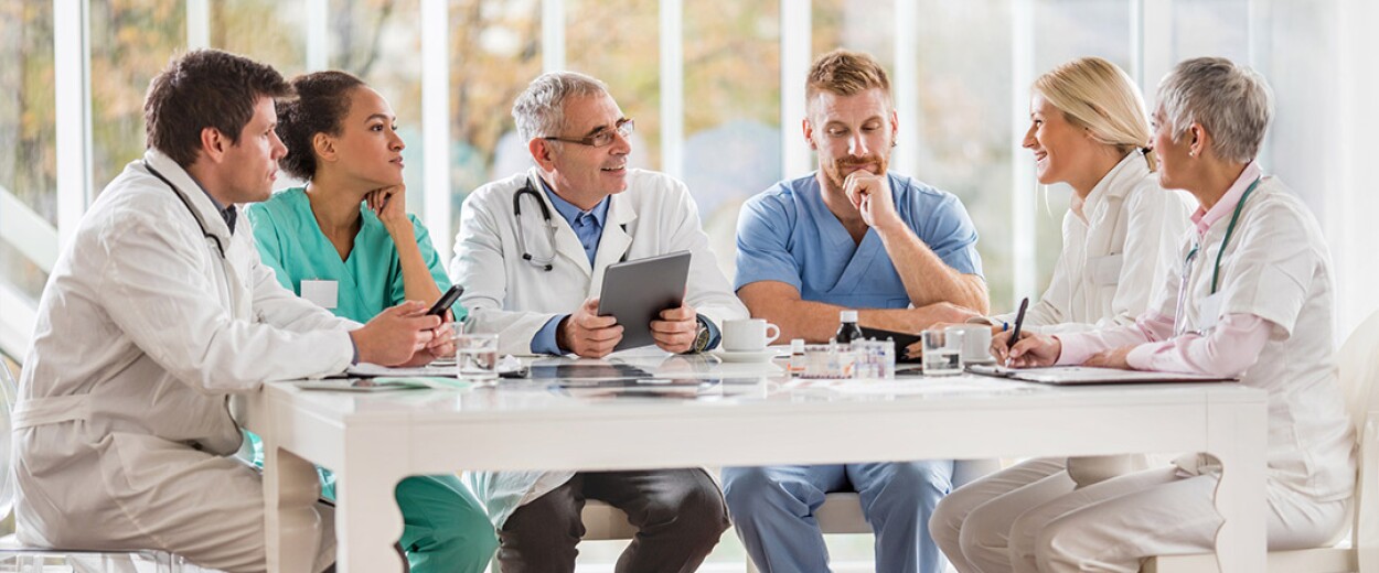 A group of medical personnel sitting at a white table talking, smiling, and taking notes