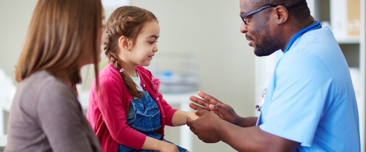 Male nurse in scrubs smiling and talking to woman and her young daughter in examination room setting