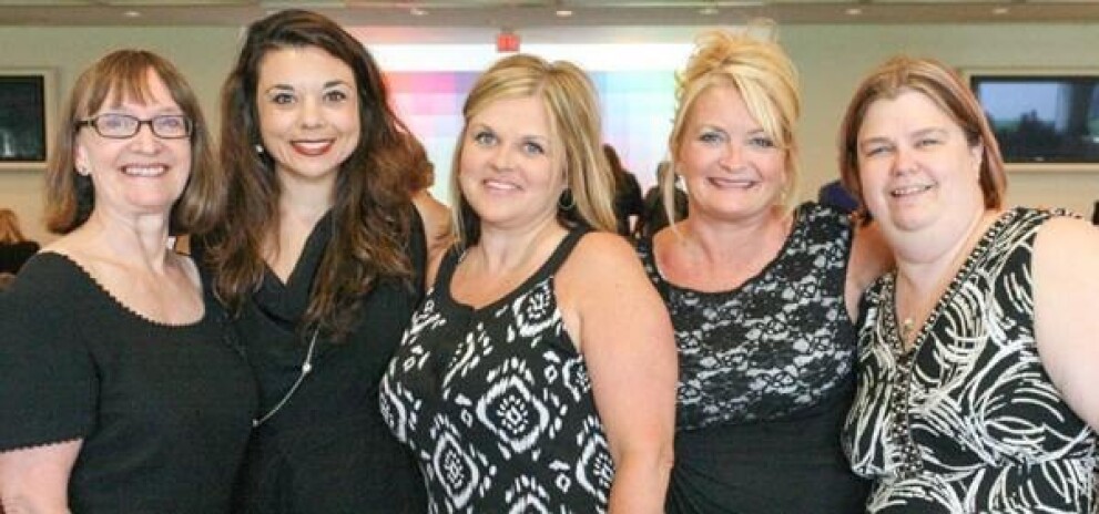 A group of 5 woman with their arms around each other smiling at the camera