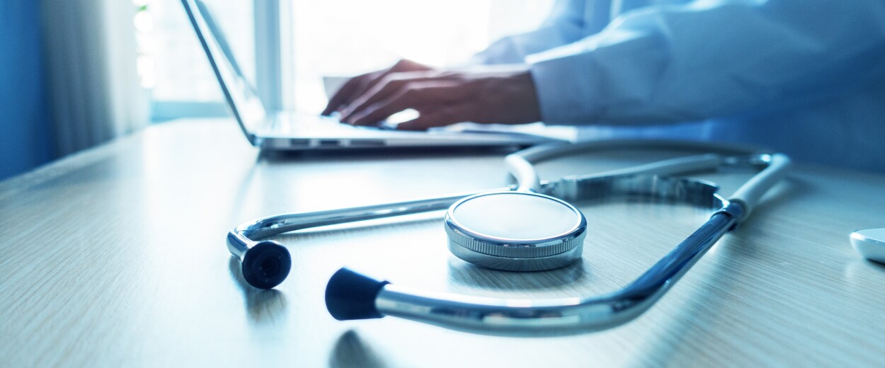 Image of stethoscope on a table next to a person in a lab coat typing on a laptop