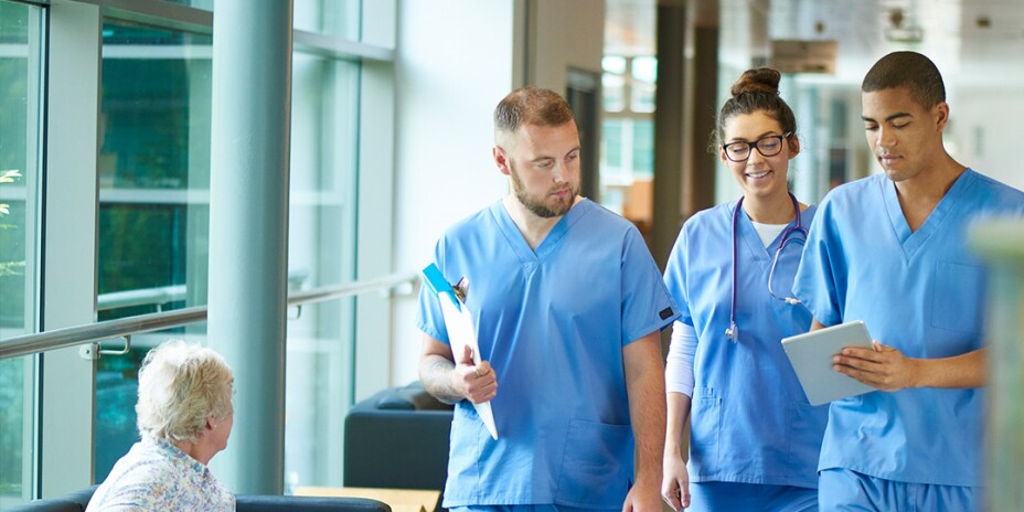Three nurses in scrubs looking at a tablet walking down a hospital corridor