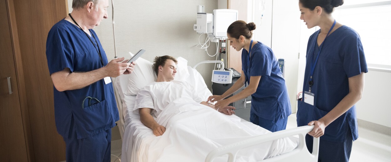 Image of a male nurse and two female nurses in scrubs checking the vitals of a male patient in hospital bed