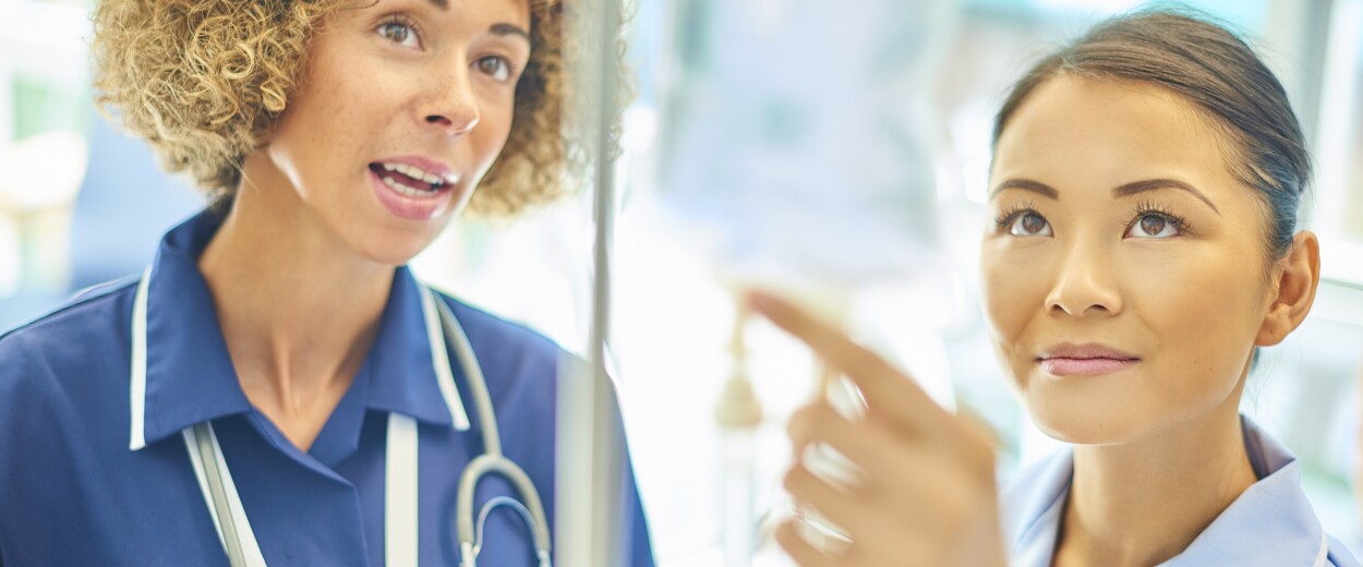 Two female nurses in scrubs looking at an IV drip and talking